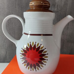 Könnte beinhalten: Eine weiße Keramik-Teekanne mit braunem Deckel und Henkel. Der Körper zeigt ein kreisförmiges Blumenmuster in Rot, Braun und Gold. Die Teekanne steht auf einem orangefarbenen Buch mit dem in Weiß gedruckten Text DA LOUISIANA STJAL BILLEDET.