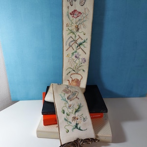 Peut inclure: Deux tentures murales brodées vintage. La plus longue représente une charrette, des fleurs et des outils de jardinage. La plus courte affiche une théière, des fleurs et un support en métal décoratif. Les deux sont sur un tissu crème avec une bordure marron.