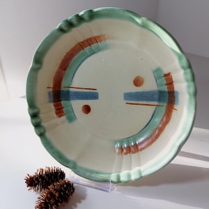 Może przedstawiać: Biały talerz ceramiczny z zielonym rantem i geometrycznym wzorem w kolorze brązowym, niebieskim i zielonym. Wzór zawiera dwa koła i dwie poziome linie.