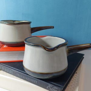 Può includere: Due brocche per salsa in ceramica color crema con manici marroni e bordi blu. Le brocche sono appoggiate su libri con copertine arancioni, blu e bianche. Le brocche hanno un design unico con bordo ondulato.
