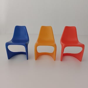 Könnte beinhalten: Drei Plastikstühle in Blau, Gelb und Orange. Die Stühle haben ein modernes, minimalistisches Design mit einer geschwungenen Rückenlehne und einer einzigen, offenen Schlaufe für einen Sitz.