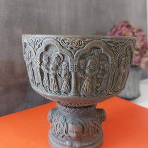 Puede incluir: Un cuenco decorativo de color bronce con intrincadas tallas en relieve que representan figuras en marcos arqueados. El cuenco se asienta sobre una base de pedestal con diseños detallados adicionales. El estilo general sugiere una estética histórica o antigua.