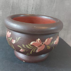 May include: A brown ceramic planter with a wide rim and a painted floral design. The flowers are pink and white with green leaves.