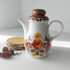 Könnte beinhalten: Weiße Keramik-Teekanne mit braunem Deckel und floralem Muster in Orange-, Gelb- und Grüntönen.
