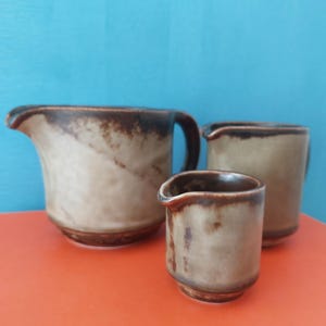 Puede incluir: Tres jarras de cerámica de diferentes tamaños. La más grande tiene asa y boquilla, las otras dos son más pequeñas. Todas son de color beige con detalles marrones y están sobre una superficie naranja sobre un fondo azul.