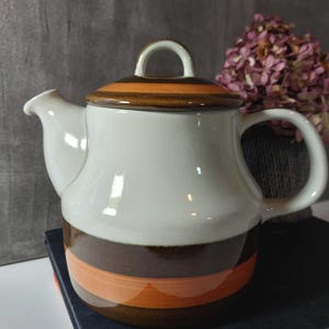 Op de afbeelding: Witte keramische theepot met een bruin en oranje gestreept ontwerp. De theepot heeft een gebogen tuit, een handvat en een deksel met een lusvormige handgreep. De theepot staat op een donkere ondergrond.
