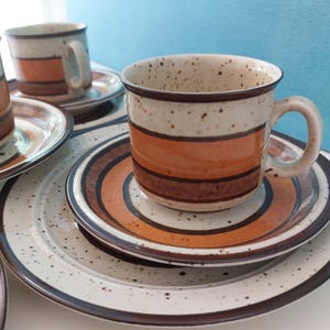 Puede incluir: Un juego de tazas y platillos de café de cerámica vintage. Las tazas y los platillos tienen un fondo crema moteado con rayas horizontales marrones y naranjas. Los bordes son marrones.