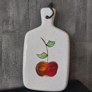 Può includere: Tagliere in ceramica bianca con un design di mela rossa e arancione e un gambo e foglie verdi. Il tagliere ha un foro nella parte superiore per appenderlo.