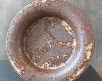 May include: A round ceramic bowl with a brown and beige glaze. The bowl has a slightly raised rim and a textured surface.