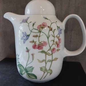 May include: White ceramic teapot with a floral design featuring pink, blue, and green flowers and foliage. The teapot has a curved handle, a spout, and a lid. The teapot is sitting on a dark surface.