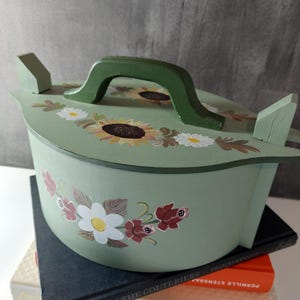 May include: A light green wooden container with a lid, featuring hand-painted sunflowers and floral designs. The container has a dark green handle and side accents. It rests on a stack of books.