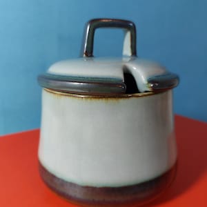 Puede incluir: Un azucarero de cerámica con tapa y asa. El cuenco es azul claro con una base y borde marrón oscuro. La tapa tiene un asa y una ranura para una cuchara. El fondo es azul y naranja.