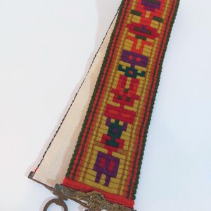 Könnte beinhalten: Ein gewebtes Lesezeichen aus Stoff mit einem geometrischen Muster in Rot-, Orange-, Grün- und Violetttönen. Das Lesezeichen hat einen Metallverschluss mit einem dekorativen Design.