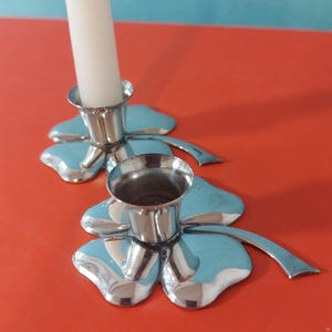 Könnte beinhalten: Zwei silberfarbene Kerzenhalter mit Kleeblatt-Design. Einer hält eine weiße Kerze. Die Kerzenhalter stehen auf einer roten Oberfläche. Die Kleeblätter haben einen hellblauen Farbton.