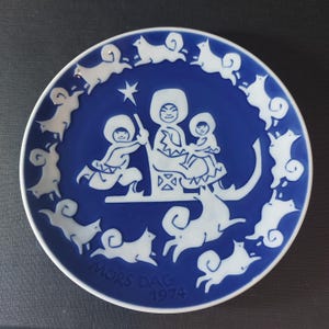 Könnte beinhalten: Ein blauer und weißer Porzellanteller mit einem Design einer Frau und zwei Kindern, die auf einem von Hunden gezogenen Schlitten fahren. Der Teller trägt den Text "MORS DAG 1974" auf der Unterseite.