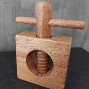 May include: A wooden nutcracker with a square base and a cylindrical handle. The handle has a crossbar for leverage. The nutcracker is made of light brown wood and has a threaded mechanism visible in a circular opening.