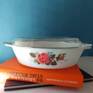 Op de afbeelding: Een witte glazen ovenschaal met een doorzichtig glazen deksel, voorzien van een bloemmotief van rode rozen en groene bladeren aan de zijkant. De schaal heeft geïntegreerde handgrepen aan beide zijden en staat op twee gestapelde boeken met oranje en zwarte kaften.