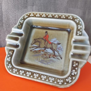 Può includere: Un posacenere quadrato in ceramica verde e marrone con un bordo a trifoglio. Il centro presenta una scena di caccia con un uomo a cavallo e due cani.