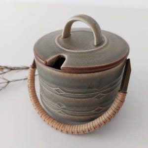 Può includere: Una zuccheriera in ceramica con coperchio e manico, con un manico intrecciato e una piccola apertura per un cucchiaio. La ciotola è di colore verde tenue con un bordo marrone e linee decorative incise.