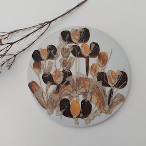 Può includere: Un piatto da parete rotondo in ceramica con un design floreale marrone e giallo. Il piatto presenta fiori stilizzati con steli marroni e centri gialli.