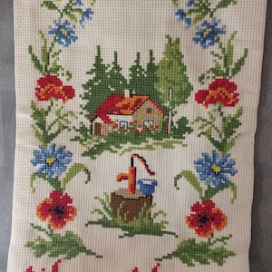 Può includere: Arazzo a punto croce con una casa, alberi e fiori. Il disegno comprende una casa con il tetto rosso, alberi verdi e fiori colorati. Il testo "til vort hjem" è ricamato in rosso in basso.