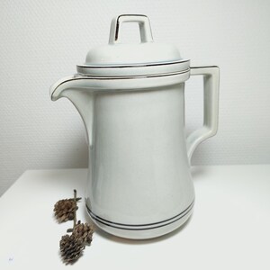 May include: A white ceramic creamer with a lid and a gold trim. The creamer has a spout and a handle. The creamer is sitting on a white surface.