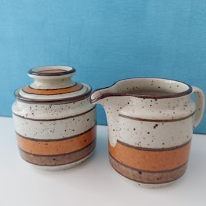 Puede incluir: Un azucarero de cerámica con tapa y una jarra para crema. El conjunto presenta un fondo beige moteado con bandas horizontales naranjas y marrones. La jarra para crema tiene un asa y un pico. El azucarero es redondo.