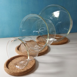 Könnte beinhalten: Klare Glaskuppeln für Terrarien in verschiedenen Größen, die auf runden Korkunterlagen ruhen. Die Kuppeln sind kaskadenartig angeordnet, mit einem blauen Hintergrund und einer weißen Oberfläche.