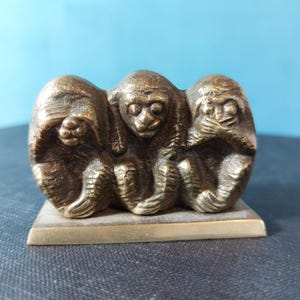 Puede incluir: Una escultura de color bronce de los tres monos sabios: no ver el mal, no oír el mal, no hablar el mal. Los monos están colocados uno al lado del otro sobre una base rectangular. La escultura tiene una textura detallada.