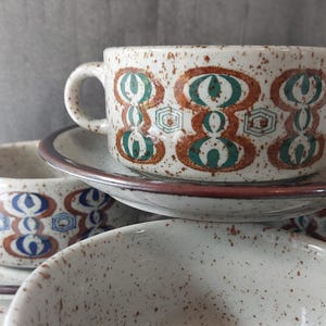 May include: A set of ceramic soup bowls and saucers featuring a speckled cream base with brown and teal geometric patterns. The bowls have a rounded handle and a wide rim, while the saucers have a dark brown edge. The design includes repeating oval shapes and small hexagonal accents.