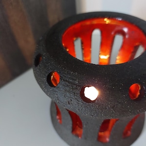 May include: A black ceramic candle holder with a textured surface and circular cutouts. The interior is a vibrant red, and a lit candle glows within, creating a warm ambiance. The design is modern and artistic.