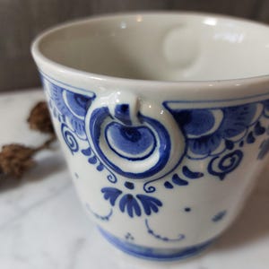 Może przedstawiać: Biały ceramiczny kubek z niebieskim kwiatowym wzorem i uchwytem. Kubek ma szczegółowy niebieski wzór wokół krawędzi i na korpusie. Uchwyt jest również ozdobiony niebieskimi akcentami. Kubek jest prawdopodobnie do picia napojów.