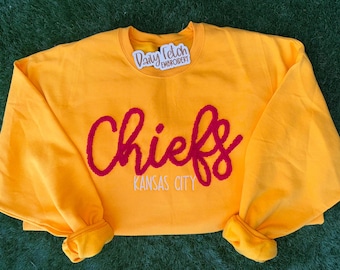 May include: A golden yellow sweatshirt with the word "Chiefs" in red script and "Kansas City" in white block letters. The sweatshirt has a crew neck and long sleeves with ribbed cuffs. A small tag is visible.