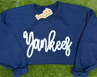 May include: Navy blue sweatshirt with the word "Yankees" in white, textured lettering. The sweatshirt has long sleeves and a crew neck. A small tag that says "Daisy Fetch Embroidery" is attached to the collar.
