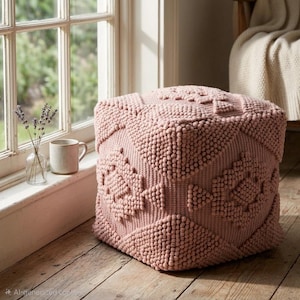 Op de afbeelding: Een roze, kubusvormige poef met een textuur, verhoogd stippenpatroon en geometrische ontwerpen. De poef is gemaakt van een zacht, pluche materiaal en staat op een houten vloer. Een mok en vaas met lavendel zijn zichtbaar.