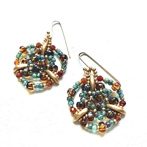 May include: A pair of handmade earrings featuring a cluster of colorful beads in shades of teal, brown, and gold. The beads are arranged in a circular pattern and attached to gold-colored metal accents and ear wires. The earrings are displayed on a white surface.