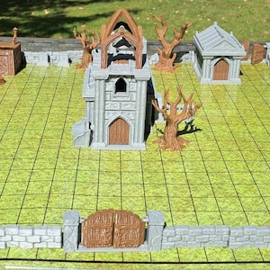May include: A miniature fantasy graveyard scene with a stone wall, gate, and buildings. The scene includes a church, mausoleum, and trees. The base is a green grid, suggesting a tabletop game setting.