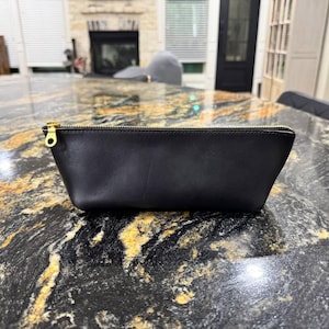 May include: A black leather zippered pouch with a gold zipper pull. The pouch is sitting on a black and gold countertop.