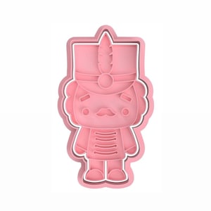 Puede incluir: Cortador de galletas de plástico rosa en forma de cascanueces de dibujos animados. El cascanueces tiene un sombrero rojo con una pluma, un bigote y un uniforme a rayas.