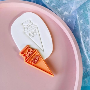 May include: A white and orange plastic cookie cutter shaped like a cone with a bow on top. The cone has the letters "ABC" printed on it.