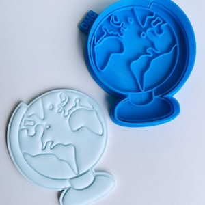 Op de afbeelding: Een blauwe plastic koekjesvormer in de vorm van een globe. De vormer heeft een reliëfdesign van de continenten en oceanen. De vormer ligt op een wit oppervlak.