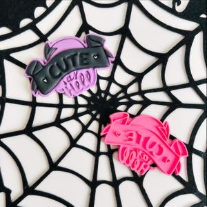 May include: Two pink and purple silicone cookie cutters shaped like banners with the text "Cute" and "Say Hello" in black lettering.