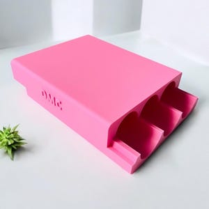 May include: A bright pink rectangular organizer with three curved slots. The word "DMC" is cut into the side of the organizer. The organizer is made of a solid, smooth material and sits on a white surface.