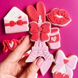 Op de afbeelding: Een set roze en rode Valentijnsdagsuikerkoekjes versierd met harten, strikken, lippen, een koffiekopje en de Eiffeltoren. De koekjes zijn gerangschikt op een roze achtergrond.