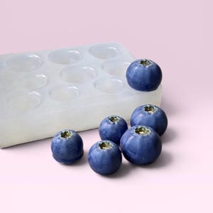 Könnte beinhalten: Nahaufnahme von mehreren frischen Blaubeeren neben einer transparenten Silikonform mit kreisförmigen Hohlräumen. Die Blaubeeren sind tiefblau und die Form ist weiß. Der Hintergrund ist ein zartes Rosa.