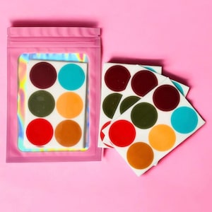 Op de afbeelding: Een roze zakje met een holografisch interieur bevat een vel met kleurrijke, cirkelvormige stickers. Extra vellen stickers worden weergegeven, met rode, bruine, groene, oranje en blauwe stippen. De achtergrond is effen roze.