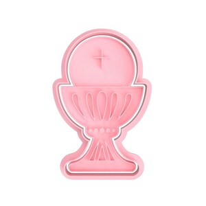 May include: Pink plastic cookie cutter shaped like a chalice with a host on top. The chalice has a cross on the host.