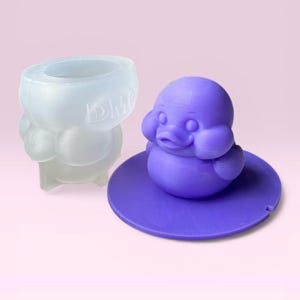 May include: A clear, duck-shaped mould and a purple duck figurine. The mould has the letters "DMC" on it. The figurine is sitting on a purple lily pad base. The duck has a round body, a beak, and small eyes.