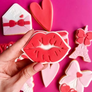 Op de afbeelding: Een set Valentijnsdag-suikerkoekjes versierd met rode, roze en witte glazuur. De koekjes hebben een hart, een strik, een lipafdruk en een Eiffeltoren.