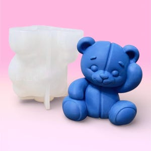 May include: A blue teddy bear figurine next to a clear, white mold in the shape of a teddy bear. The bear is sitting with one arm raised. The mold has the letters "LMC" printed on it. The background is a gradient of pink.